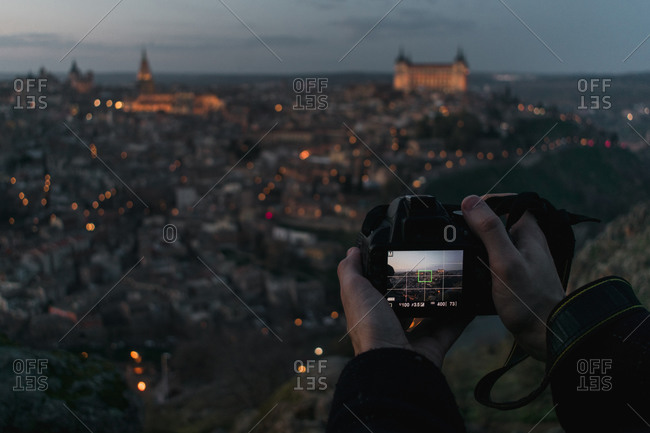 Crop hands of male photographer with camera standing on hill and taking photo of old Spanish city Toledo with castles in twilight