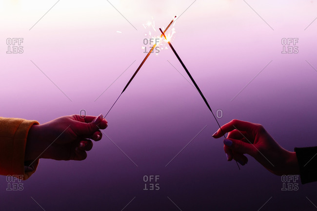 Crop female hands in front of each other holding burning sparklers on colorful purple background