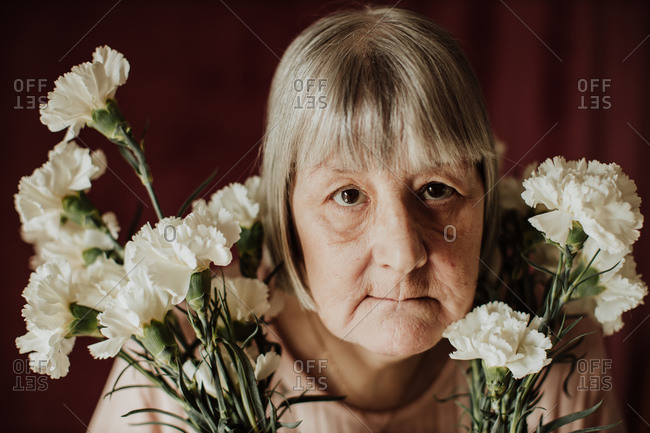 From above thoughtful old female with gray hair looking at camera holding bouquet of white carnation at home