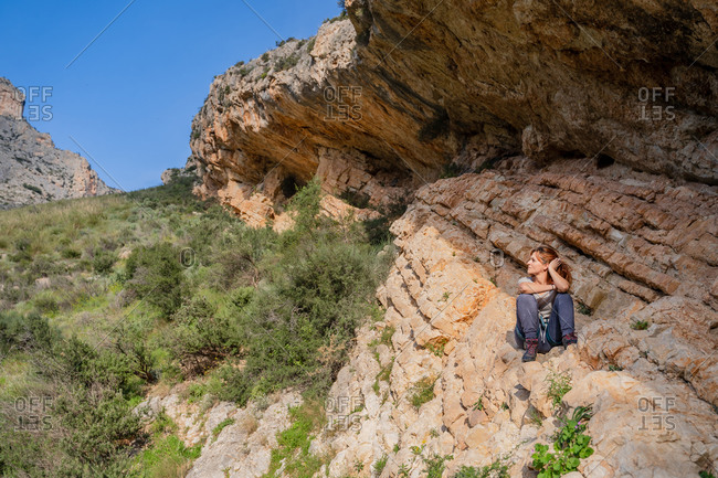 Pensive relaxed female alpinist contemplating nature while sitting under sheer cliff in summer day