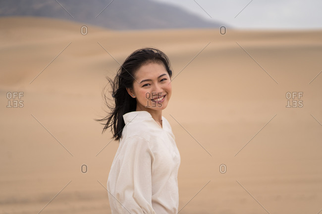 Side view of happy youthful Asian woman looking at camera while standing in middle of sandy desert in Saudi Arabia