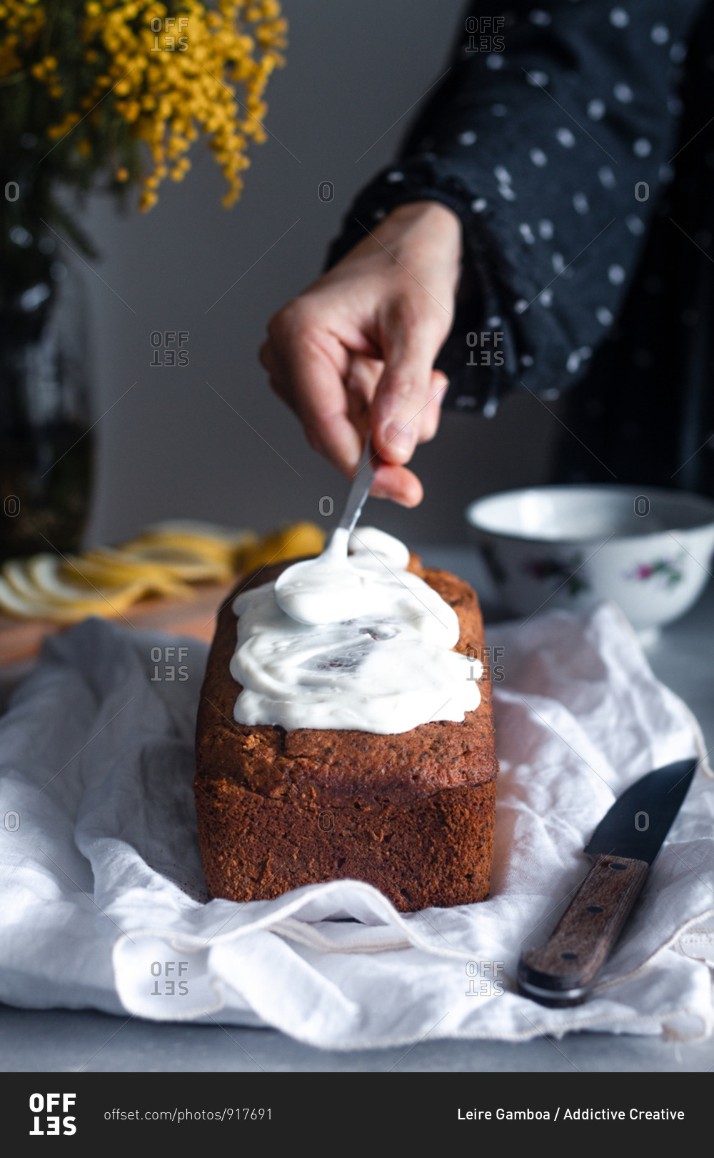 Crop hand of housewife decorating delicious homemade lemon and poppy seeds sponge cake with whipped cream at table with bouquet of mimosa flowers in background