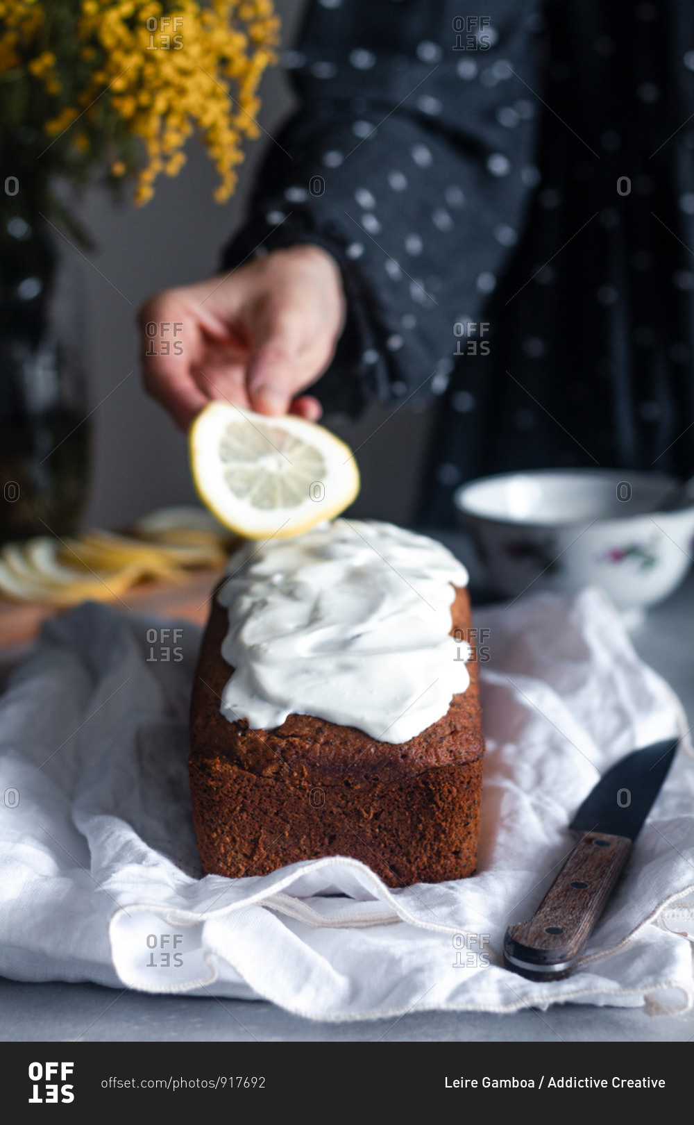Crop hand of woman putting slice of lemon on yummy fresh homemade cake covered with whipped cream placed on white cloth on kitchen table with bouquet of mimosa flowers in background