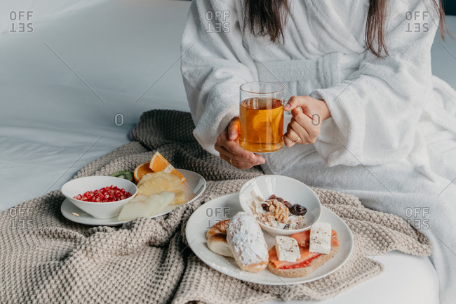 Cropped unrecognizable brunette female in white bathrobe holding glass mug of tea while having breakfast in bed with fruits and pastry