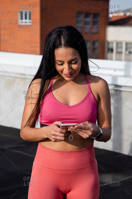 Young satisfied brunette in stylish pink bra and leggings smiling while surfing mobile phone during workout on roof of building