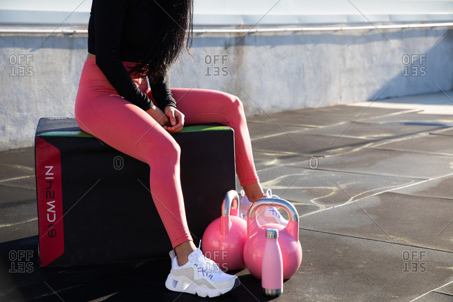 Crop legs of female athlete in stylish pink leggings and white sneakers sitting on box with dumbbells and bottle on roof of building