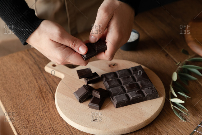 From above of crop hands of housewife breaking chocolate bar over wooden cutting board while preparing dessert at wooden table in home kitchen
