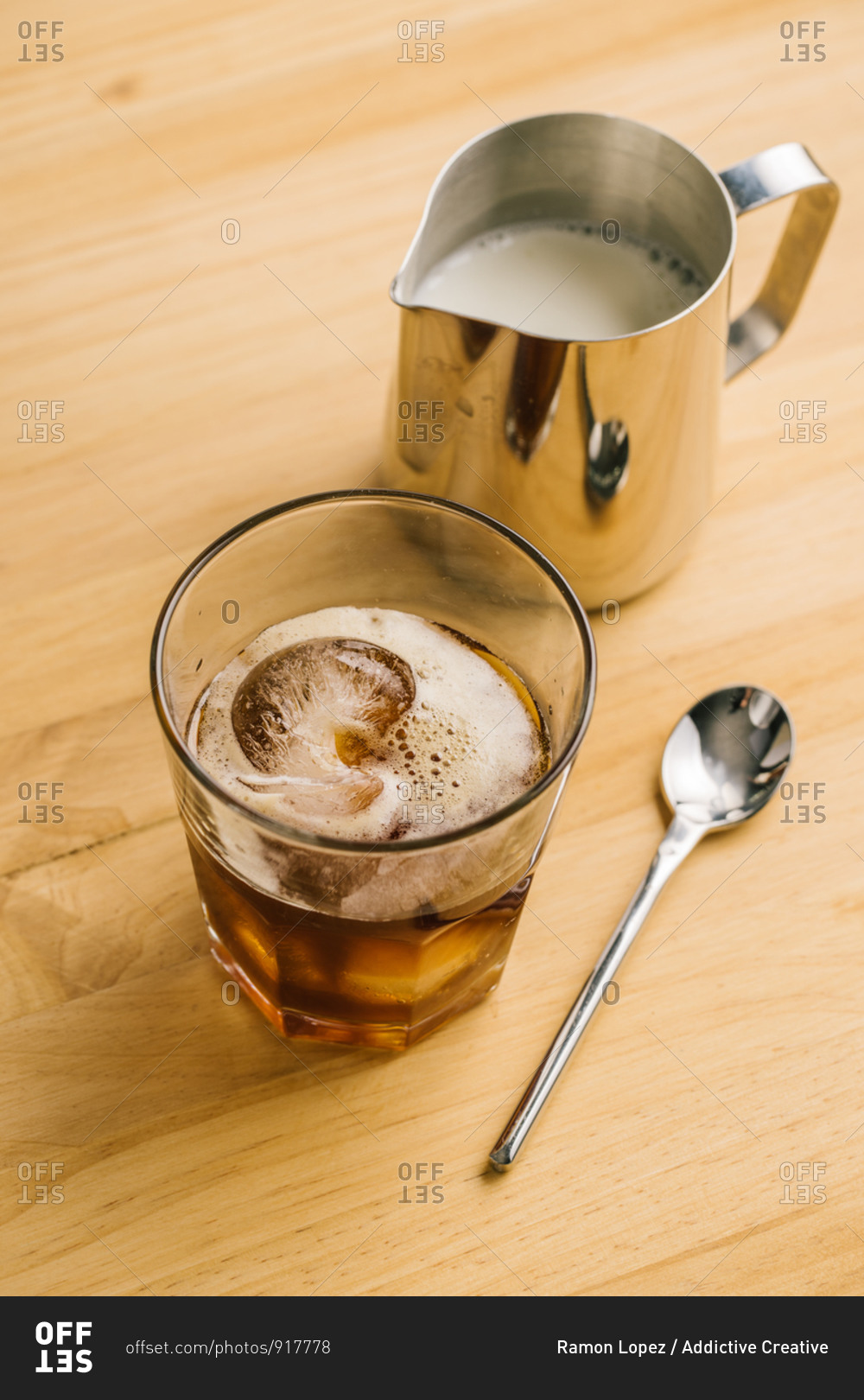 From above of stylish glass with ice black coffee on wooden table with milk pitcher and spoon