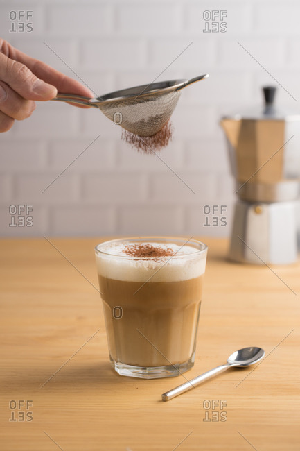 Anonymous barista sifting cacao powder by sieve on glass with brewed coffee and milk foam in cafe