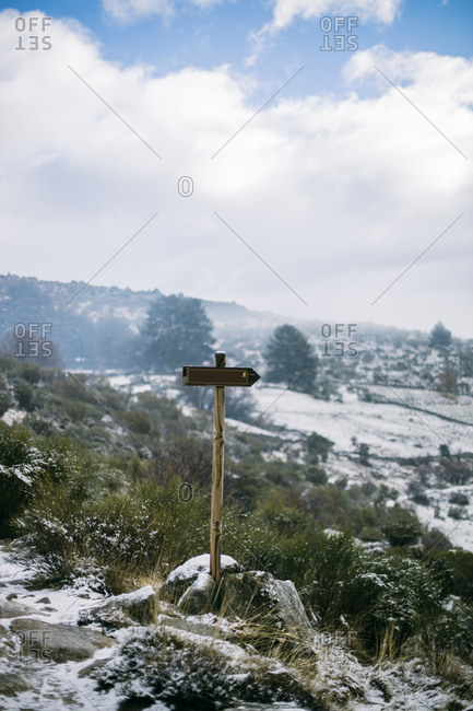 Small wooden guide sign on rocky roadside with mountainous terrain and blue cloudy sky on background