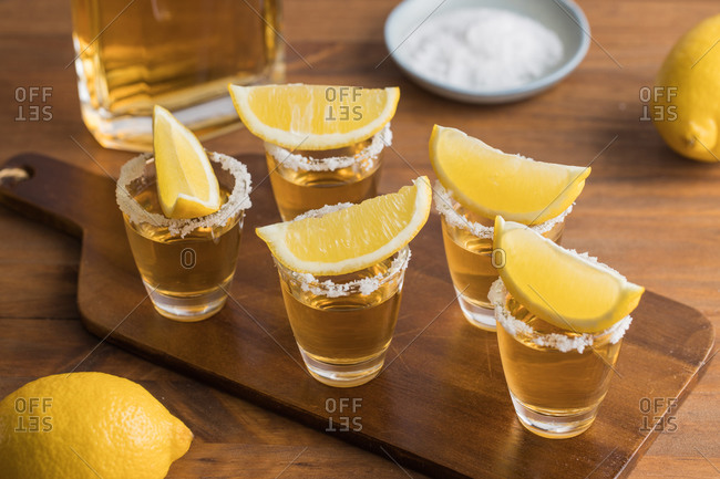 From above glass shots of golden tequila with salty rim and slices of lemon on top on wooden table