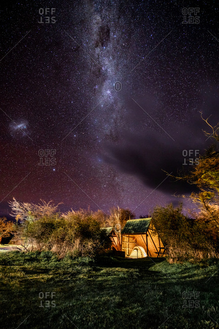 Magnificent scenery with solitary camp on glade among trees under violet clear sky with Milky Way among much of stars