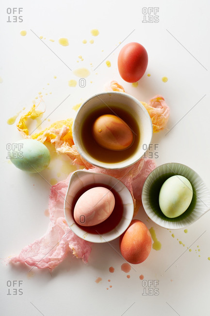 Top view image of colorful painted eggs in small white bowls on neutral background with negative space