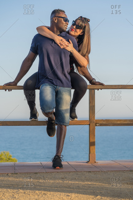 Cheerful woman smiling and hugging black boyfriend while sitting on railing against sea and sundown sky during romantic date in evening