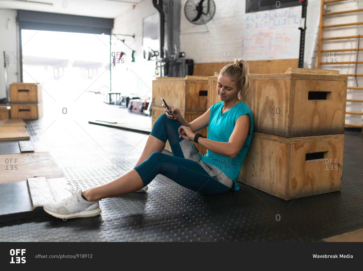 Side view of an athletic Caucasian woman wearing sports clothes cross training at a gym, taking a break from training sitting and leaning on a box, using a smartphone and smiling