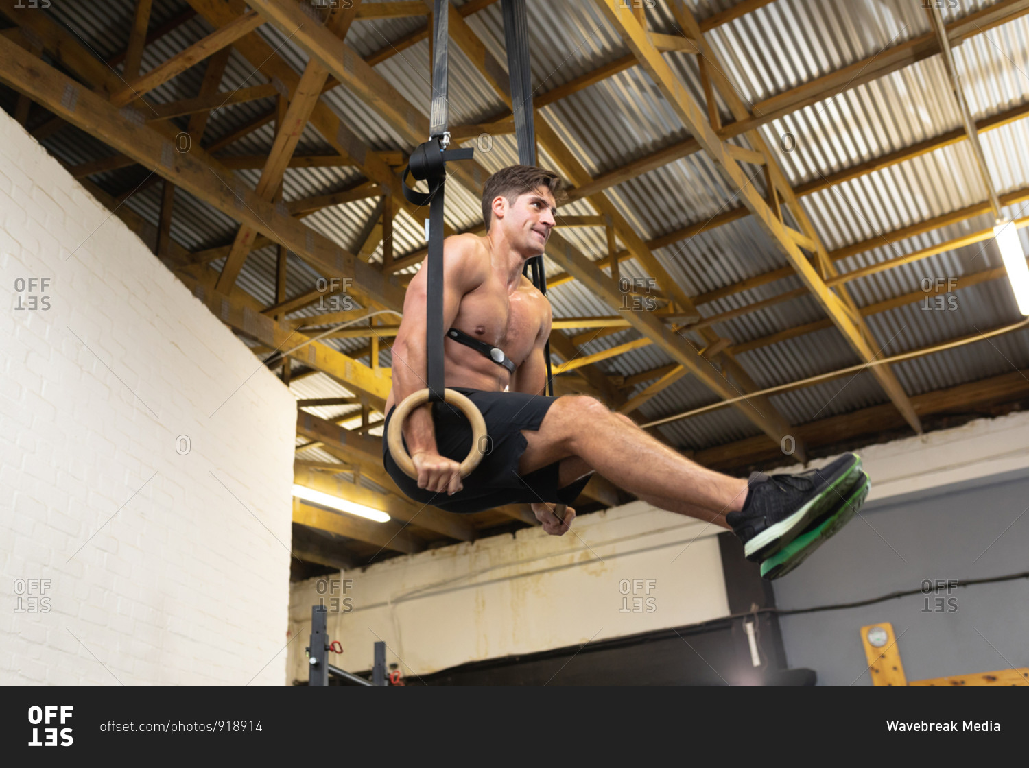 Side view of a shirtless athletic Caucasian man wearing a chest strap heart rate monitor cross training at a gym, lifting himself up on gymnastic rings with legs up