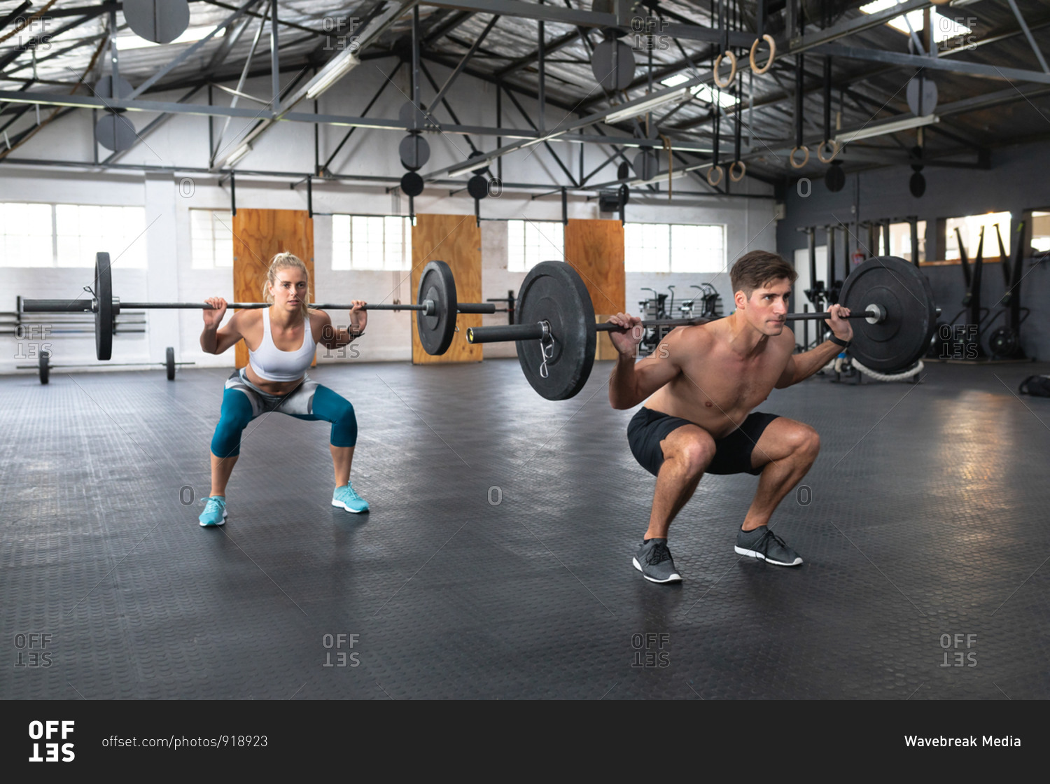 Side view of an athletic Caucasian man and woman wearing sports clothes cross training at a gym, weight training with barbells, squatting with the weights on their shoulders