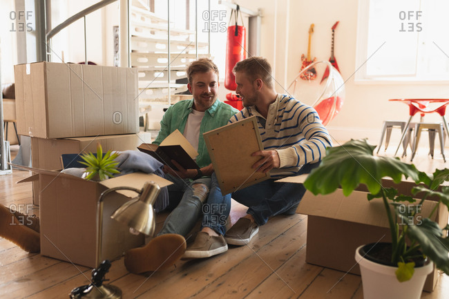 Front view of a Caucasian male couple moving in to a new apartment, sitting on the floor with cardboard boxes and interacting