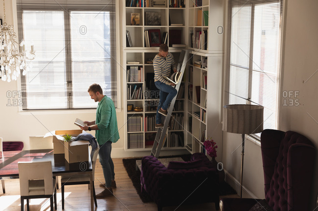 Side view of a Caucasian male couple moving in to a new apartment, unpacking their belongings from cardboard boxes and putting them on shelves, standing on a ladder.