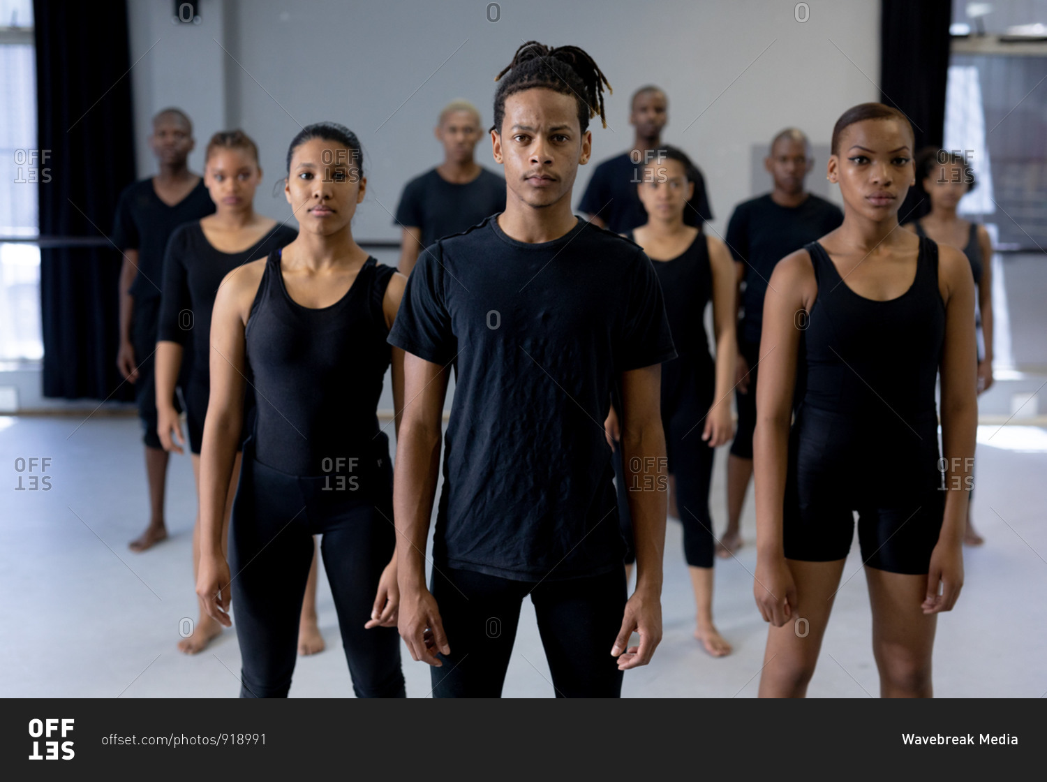 Front view of a mixed race modern male dancer wearing black clothes, standing in front of a multi-ethnic group of fit male and female dancers, looking straight into a camera.