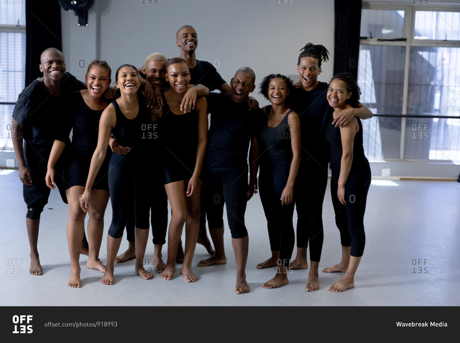 Front view of a multi-ethnic group of fit male and female modern dancers wearing black outfits practicing a dance routine during a dance class in a bright studio, standing together, smiling and looking straight into a camera.