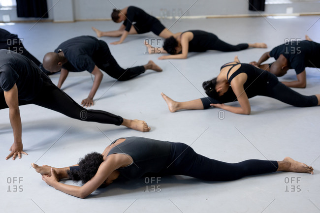 Side view of a multi-ethnic group of fit male and female modern dancers wearing black outfits practicing a dance routine during a dance class in a bright studio, lying on the floor and stretching up.