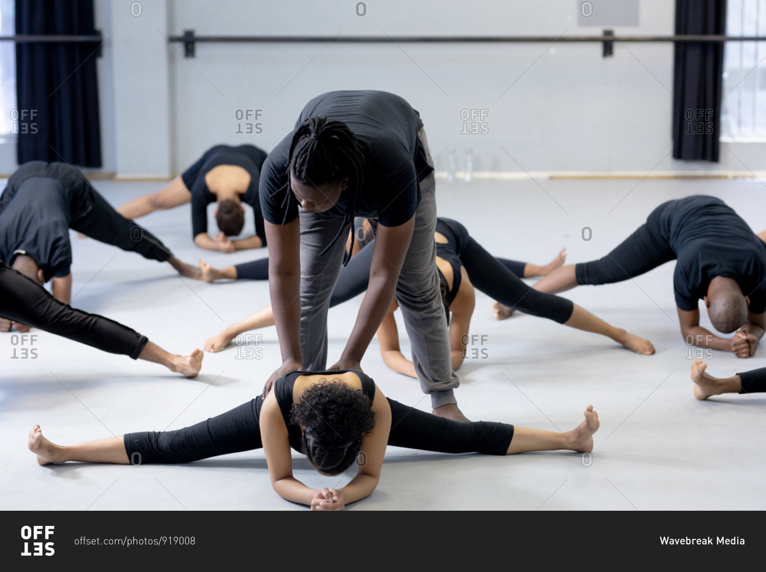 Front view of a mixed race fit male modern dancer wearing black outfit, supporting a female dancer while stretching up during a dance class in a bright studio, with other dancers exercising in the background.