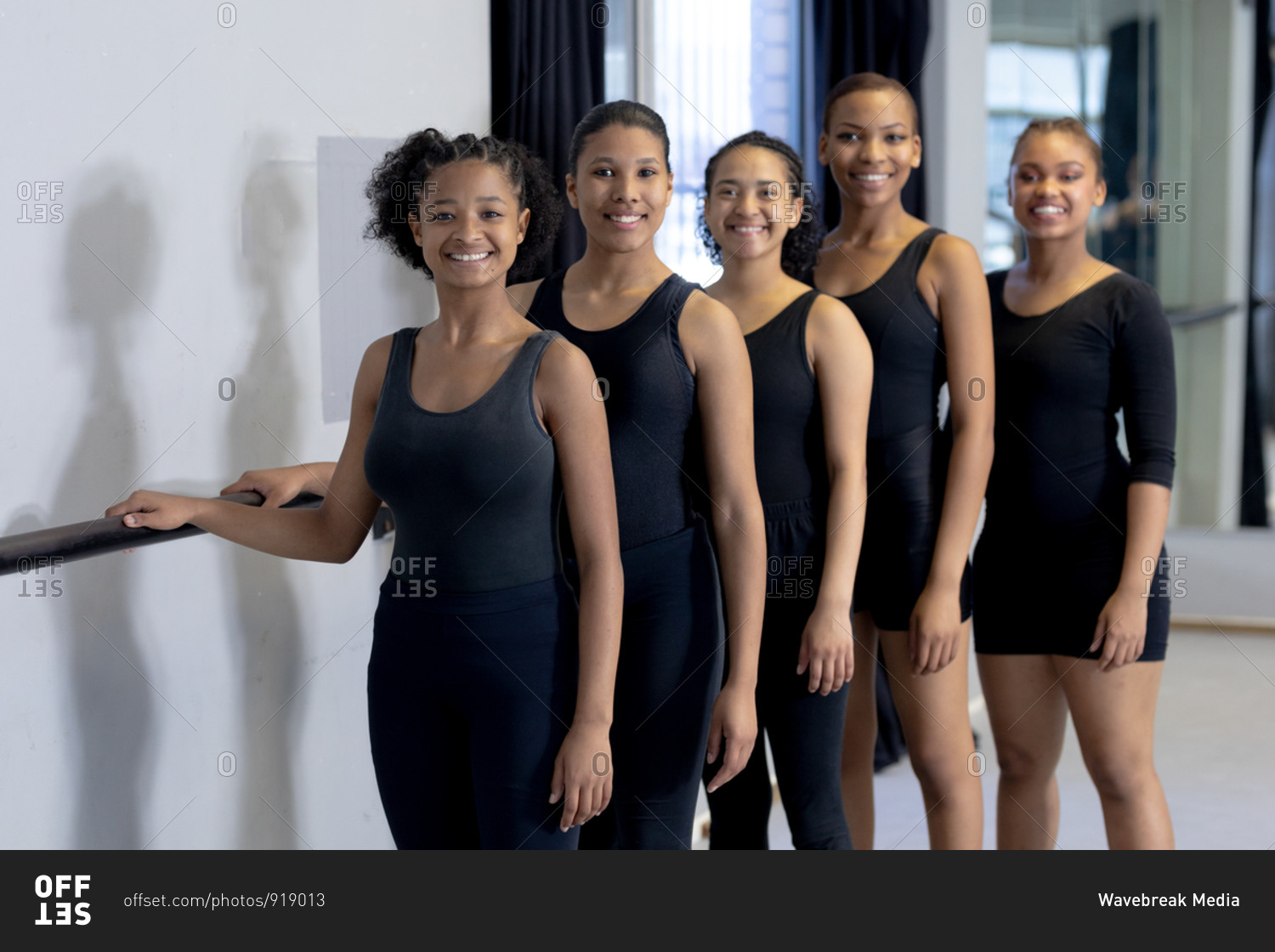 Front view of a multi-ethnic group of fit female modern dancers wearing black outfits practicing a dance routine during a dance class in a bright studio, standing by a handrail, smiling and looking straight into a camera.