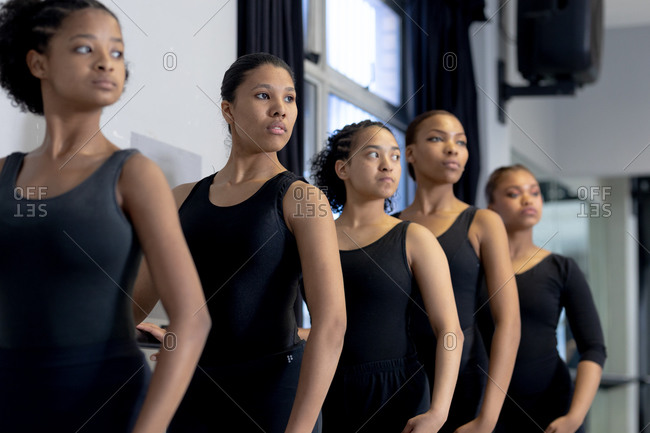 Side view close up of a multi-ethnic group of fit female modern dancers wearing black outfits practicing a dance routine during a dance class in a bright studio, standing by a handrail with their arms on their hips.