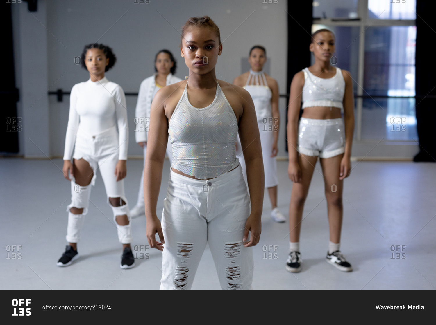 Front view of a multi-ethnic group of fit female modern dancers wearing white outfits practicing a dance routine during a dance class in a bright studio, standing on the floor and looking straight into a camera.