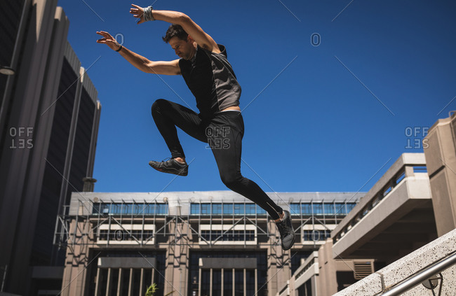 Side view of a Caucasian man practicing parkour by the building in a city on a sunny day, jumping on concrete handrail.