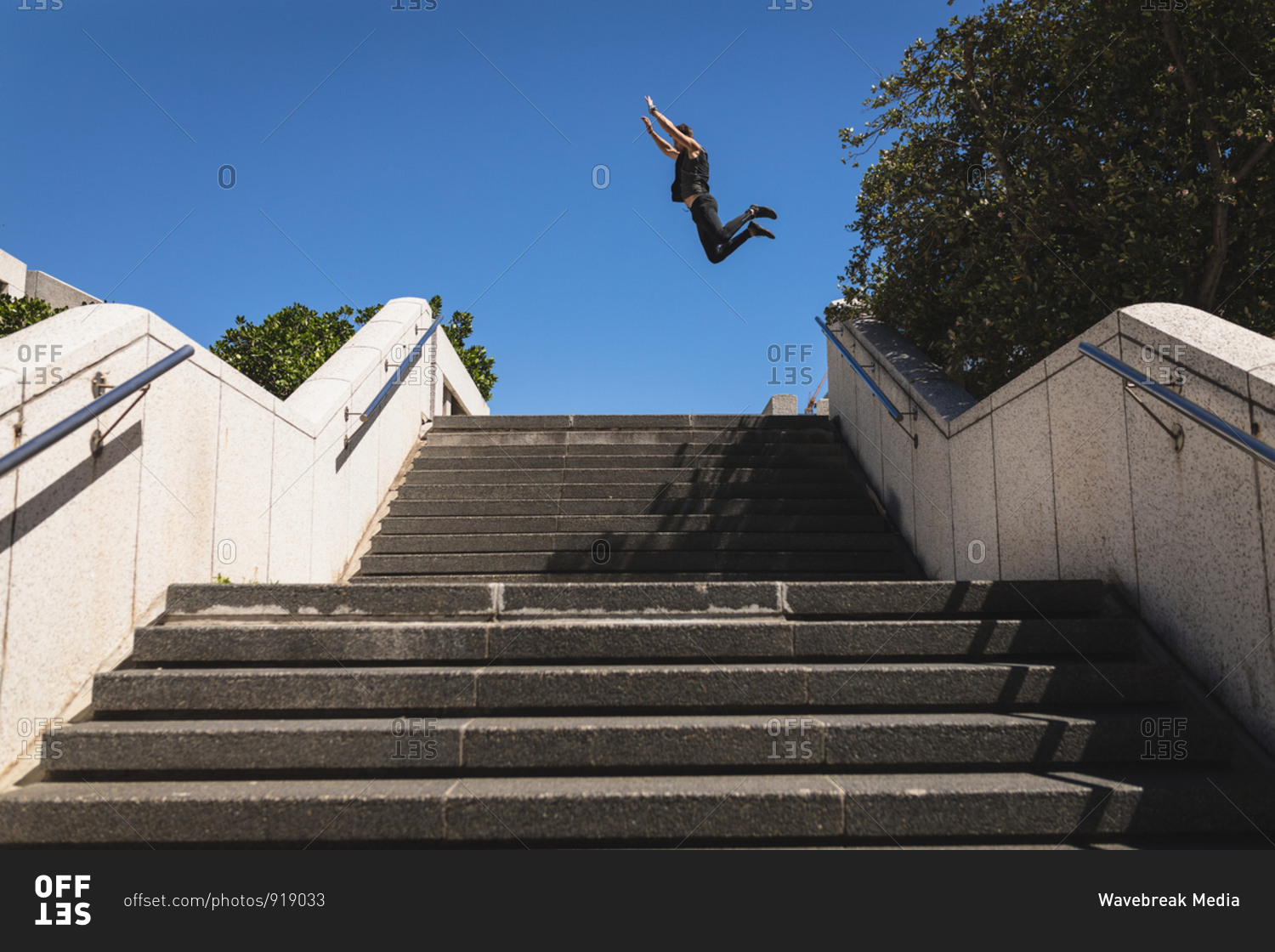 Side view of a Caucasian man practicing parkour by the building in a city on a sunny day, jumping above stairs.
