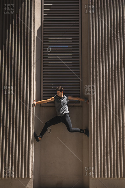 Side view of a Caucasian man practicing parkour by the building in a city on a sunny day, climbing concrete wall of a modern building.