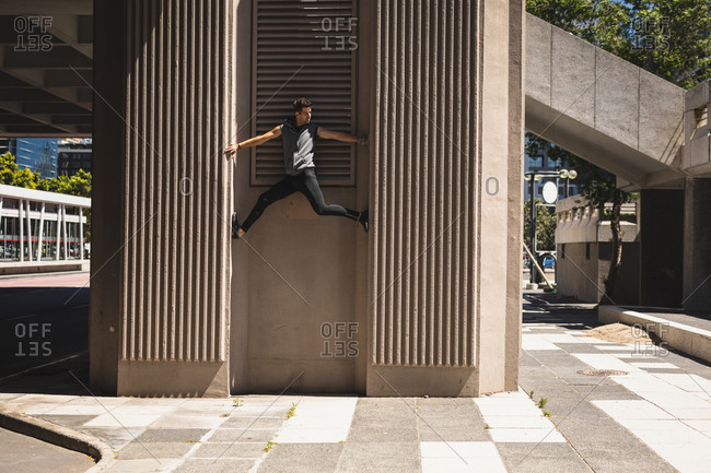 Side view of a Caucasian man practicing parkour by the building in a city on a sunny day, climbing concrete wall of a modern building.