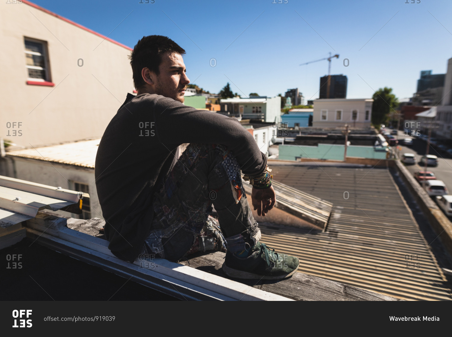 Side view of a Caucasian man practicing parkour by the building in a city on a sunny day, taking a break, resting and sitting on a rooftop.