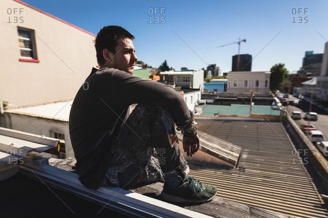 Side view of a Caucasian man practicing parkour by the building in a city on a sunny day, taking a break, resting and sitting on a rooftop.