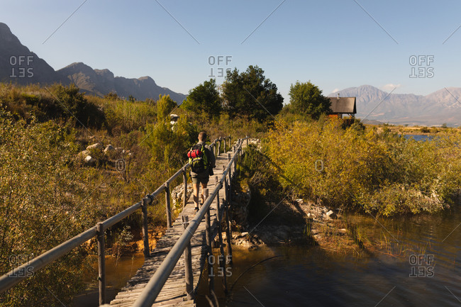 Rear view of a Caucasian man having a good time on a trip to the mountains, standing on a bridge, enjoying his view, walking towards a cabin, on a sunny day