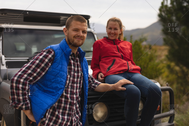Front view of a Caucasian couple having a good time on a trip to the mountains, a woman is seating on a car hood and a man is holding her thigh, looking at the camera