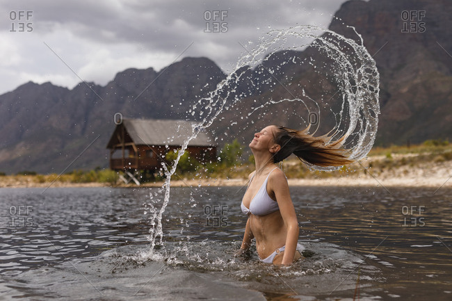 Rear view of a Caucasian woman having a good time on a trip to the mountains, standing in a lake, tossing her wet hair, leaving a water trail in the air
