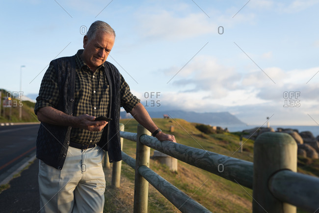 Front view of a senior Caucasian man standing alone in the countryside by the sea, using his phone, leaning on a fence by the side of a road