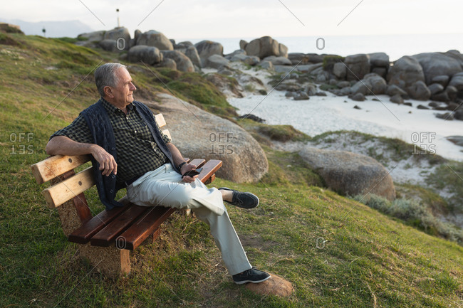 Side view of a senior Caucasian man relaxing, sitting on a bench in the countryside by the sea admiring a coastal view