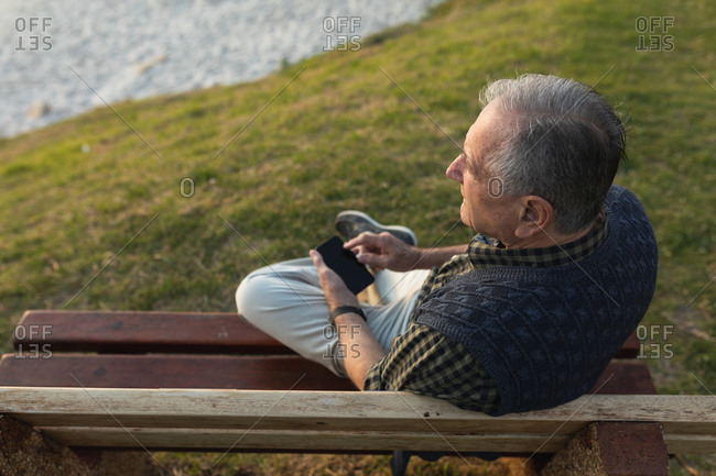 High angle view of a senior Caucasian man relaxing, sitting on a bench in the countryside by the sea admiring a coastal view and using a smartphone