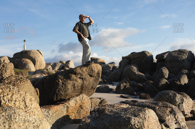 Side view of a senior Caucasian man standing alone on rocks at a beach, shielding his eyes from the sun and admiring a coastal view, with blue sky in the background