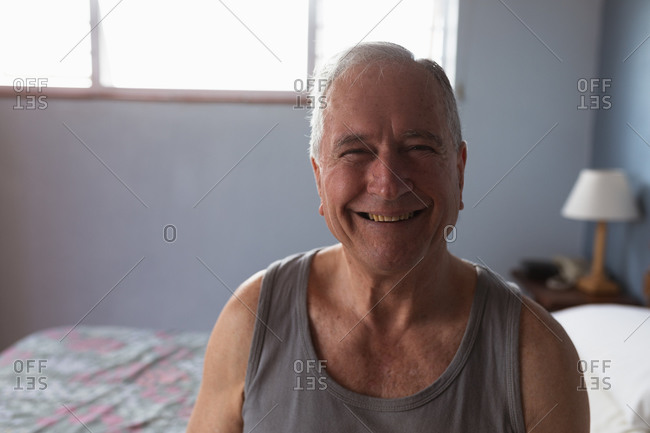 Portrait of a senior Caucasian man relaxing at home in his bedroom, wearing a vest and looking to camera smiling, with a sunlit window behind him