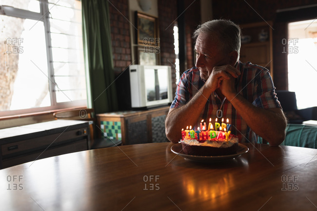 Side view of a senior Caucasian man at home, sitting alone at the dining table with a birthday cake with lit candles on it in front of him