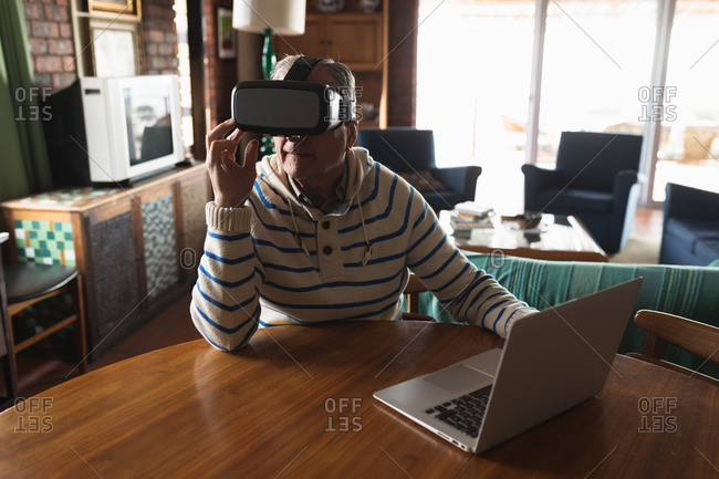 Front view of a senior Caucasian man relaxing at home in his living room, sitting at the table using a laptop computer and wearing a virtual reality headset