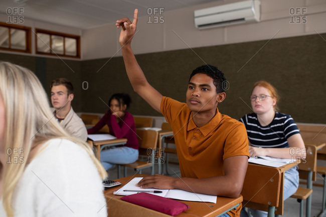 Front view of a teenage mixed-race boy in a school classroom sitting at desk, raising his hand to answer a question, with teenage male and female classmates sitting at desks working in the background