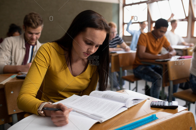 Front view close up of a teenage Caucasian girl in a school classroom sitting at desk, concentrating and reading, with teenage male and female classmates sitting at desks working in the background