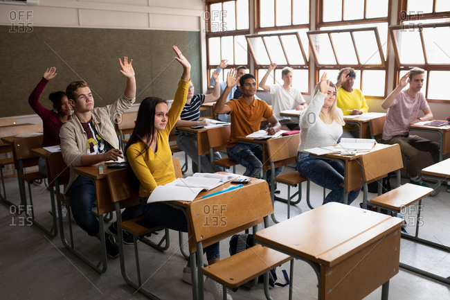 Side view of multi-ethnic group of high school teenagers in a school classroom sitting at desks, all raising their hands to answer a question