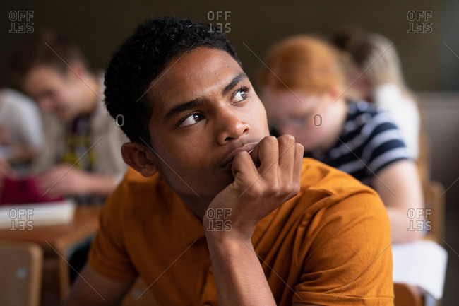 Side view close up of a teenage mimed-race boy in a school classroom sitting at desk, concentrating, with teenage male and female classmates sitting at desks working in the background
